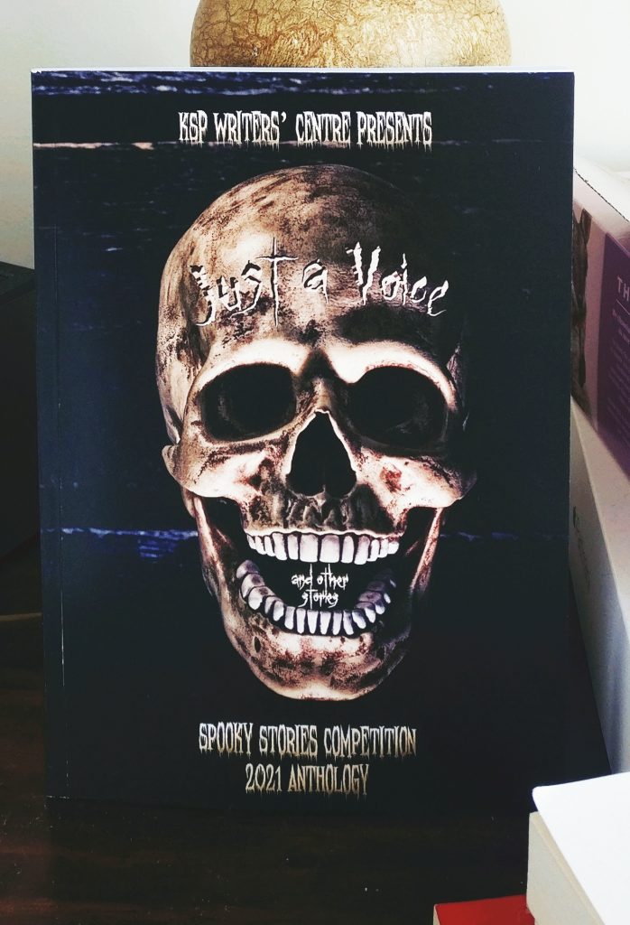 A photograph of the book cover for Just a Voice and other stories. The cover artwork has a skull on a dark background. The main title looks carved into the skull
